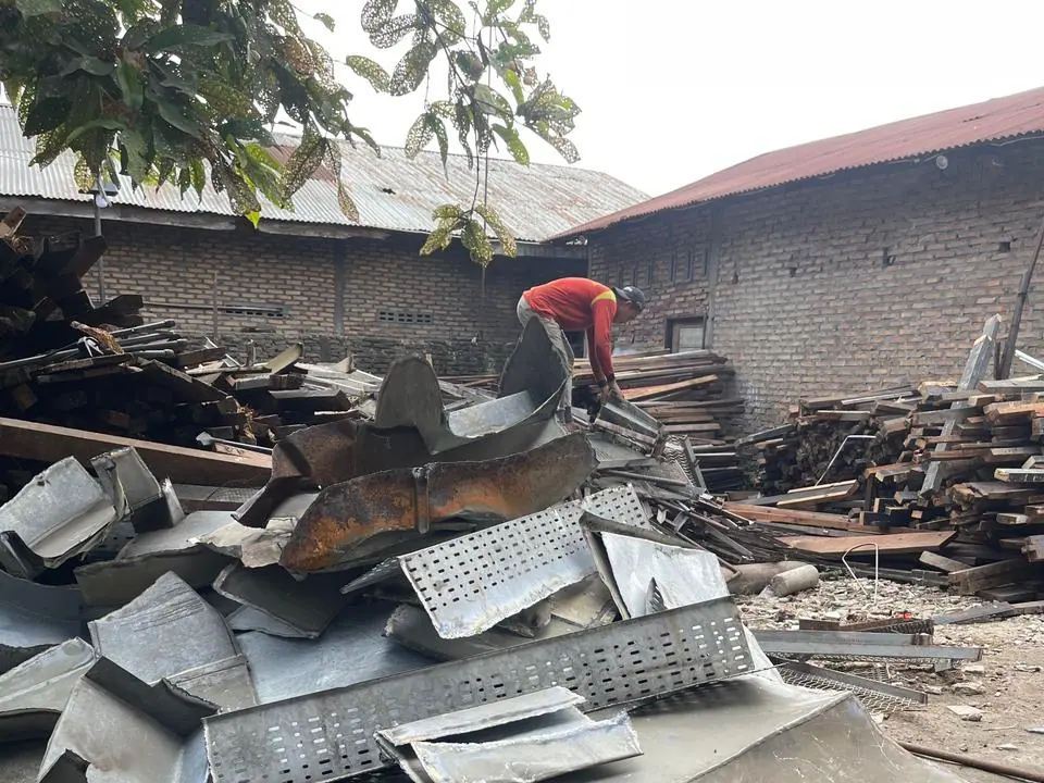 Tumpukan seng dan besi bekas hasil bongkaran dengan pekerja memilah di latar belakang